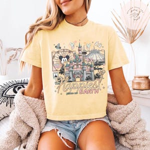 May include: Yellow t-shirt featuring a graphic of a Disney castle, Mickey Mouse, and the text "The Happiest Place on Earth." The design includes a Ferris wheel, Space Mountain, and other park elements. The shirt is a light yellow color.