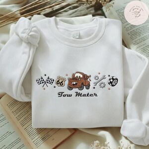 May include: A white crewneck sweatshirt featuring an embroidered design of a brown tow truck, a Route 66 sign, checkered flags, a wrench, a gear, and a tire. The text "Tow Mater" is embroidered in dark brown lettering below the truck graphic.