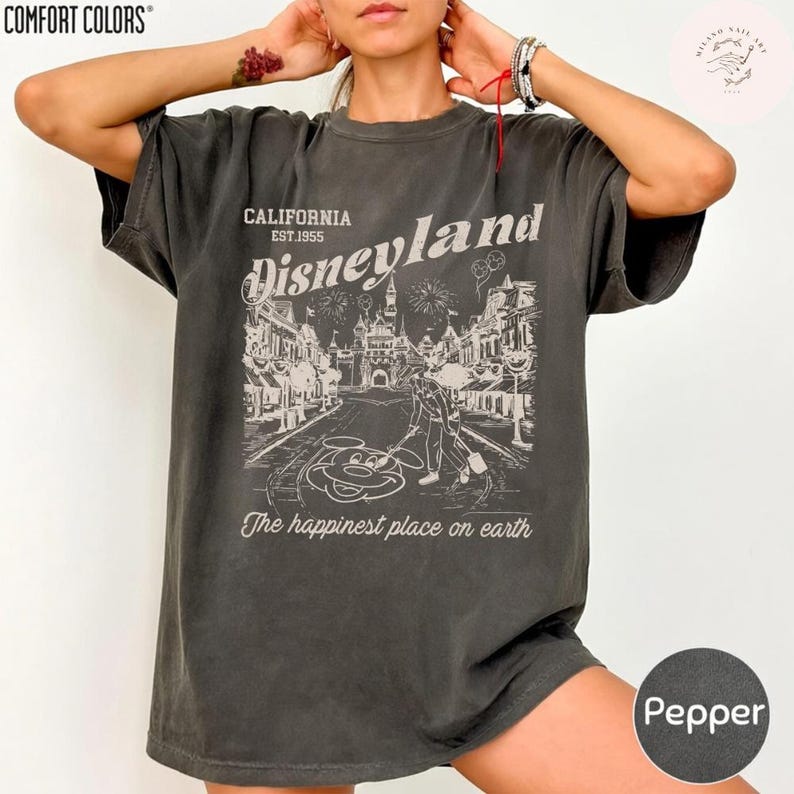 May include: Pepper-colored t-shirt with a vintage Disneyland graphic. The design features the text "CALIFORNIA EST.1955 Disneyland" above a detailed illustration of a street scene with Mickey Mouse. The phrase "The happiest place on earth" is below the graphic.