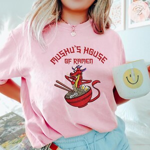 Op de afbeelding: Een lichtroze t-shirt met de tekst "Mushu's House of Ramen" in het rood. Het shirt heeft een cartoonillustratie van een rode draak in een kom ramen met eetstokjes. Een witte mok met een gele smiley is ook zichtbaar.