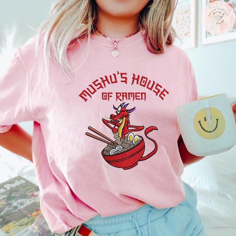Op de afbeelding: Een lichtroze t-shirt met de tekst "Mushu's House of Ramen" in het rood. Het shirt heeft een cartoonillustratie van een rode draak in een kom ramen met eetstokjes. Een witte mok met een gele smiley is ook zichtbaar.
