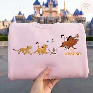 May include: A pale pink sweatshirt with an embroidered design from Disney's The Lion King. The design features Simba, Timon, Pumbaa, and a bird. The words "The Lion King" are embroidered in gold.