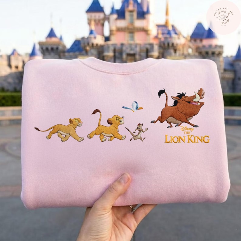 May include: A pale pink sweatshirt with an embroidered design from Disney's The Lion King. The design features Simba, Timon, Pumbaa, and a bird. The words "The Lion King" are embroidered in gold.