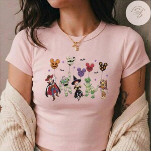 May include: A light pink short-sleeved t-shirt featuring a graphic print of five animated characters dressed in Halloween costumes, each holding a Mickey Mouse-shaped balloon. The characters include a cowboy, a space ranger, a cowgirl, a dinosaur, and a dog.