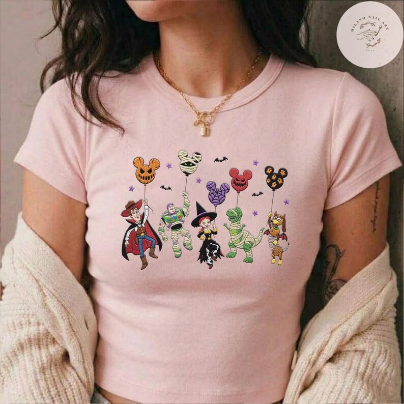 May include: A light pink short-sleeved t-shirt featuring a graphic print of five animated characters dressed in Halloween costumes, each holding a Mickey Mouse-shaped balloon. The characters include a cowboy, a space ranger, a cowgirl, a dinosaur, and a dog.