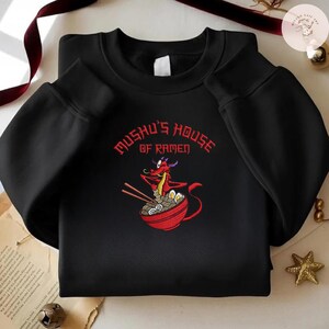 Op de afbeelding: Zwart sweatshirt met een rode cartoon draak met eetstokjes en een kom ramen. De tekst "Mushu's House of Ramen" staat in rood boven de draak. Het sweatshirt is een casual stijl.