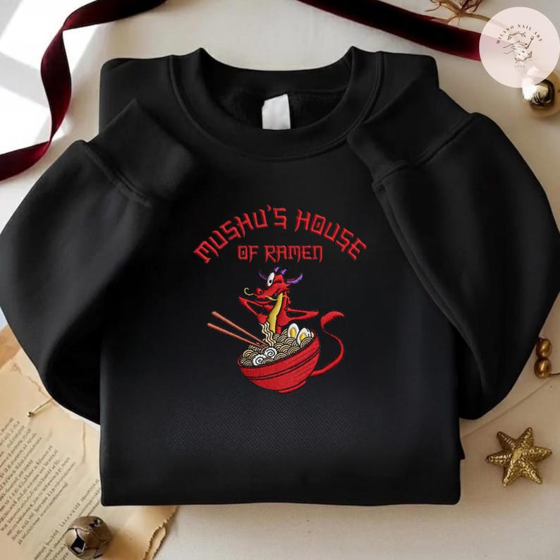 Op de afbeelding: Zwart sweatshirt met een rode cartoon draak met eetstokjes en een kom ramen. De tekst "Mushu's House of Ramen" staat in rood boven de draak. Het sweatshirt is een casual stijl.