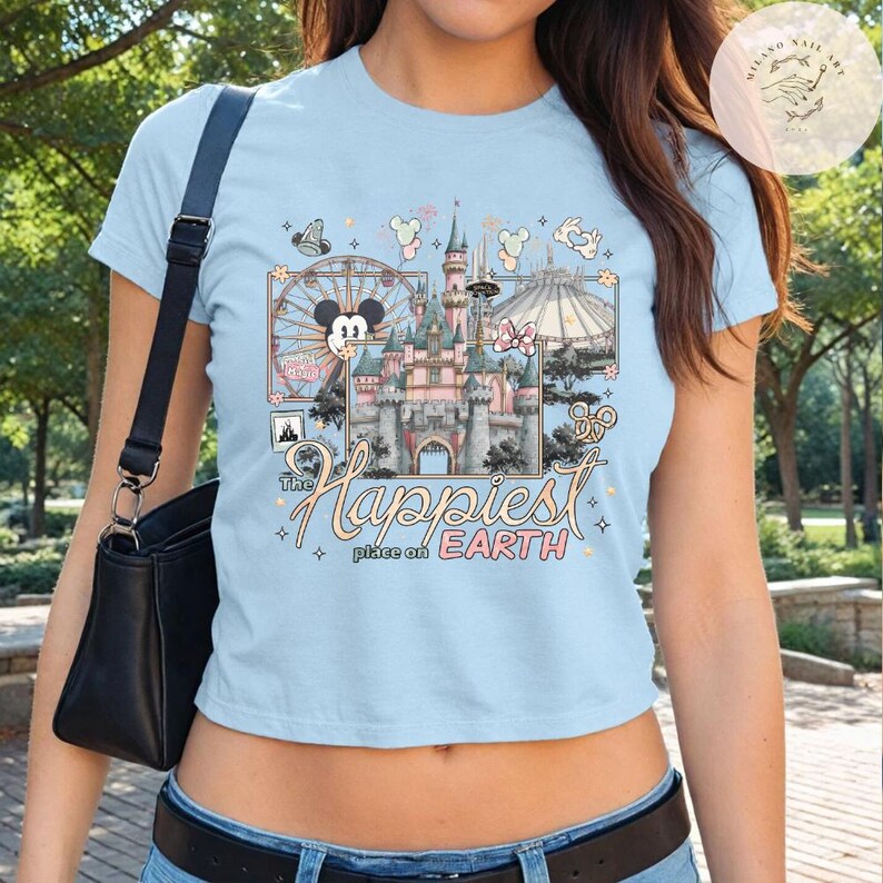 May include: Light blue t-shirt featuring a colorful graphic of a castle, a Ferris wheel, and other theme park elements. The text "The Happiest Place on Earth" is displayed in a script font. The shirt has short sleeves and a crew neck.