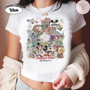 Puede incluir: Camiseta blanca con un gr&aacute;fico colorido de personajes de Disney, incluyendo Mickey Mouse, Buzz Lightyear y Ariel. El texto "Hollywood Studio Walt Disney World" est&aacute; impreso en la parte inferior. La camiseta tiene las palabras "Comfort Colors" y "White" impresas en la parte superior izquierda.