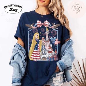 May include: Navy blue t-shirt with a patriotic design featuring a princess in a dress with an American flag pattern, a castle, fireworks, and a Mickey Mouse-shaped balloon. The shirt has the words "Comfort Colors Navy" in a white oval.
