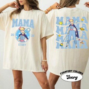 May include: Ivory-colored t-shirt with the word "MAMA" in light blue, featuring a cartoon character design. The front design includes the text "20 STORY 26". The back of the shirt repeats "MAMA" with stars, a planet, and a rocket. The shirt is labeled "Comfort Colors Ivory".