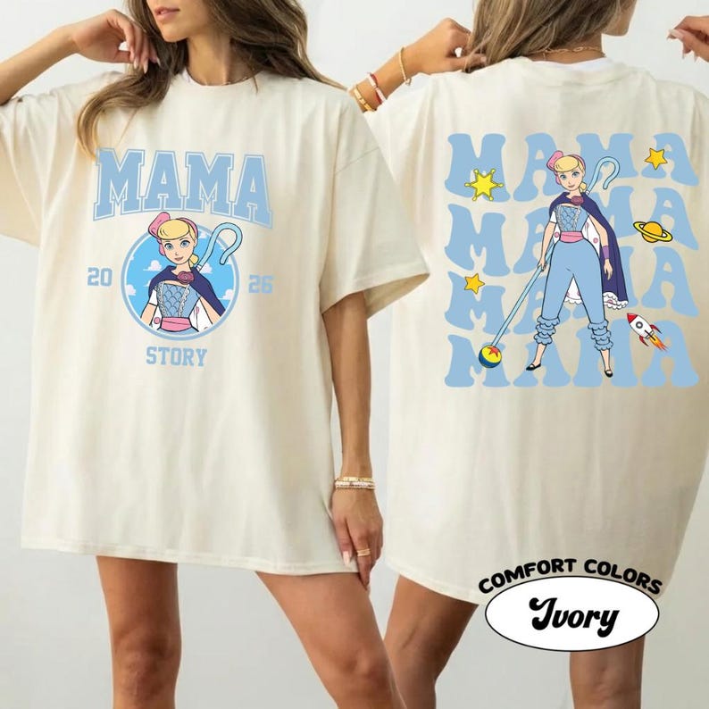 May include: Ivory-colored t-shirt with the word "MAMA" in light blue, featuring a cartoon character design. The front design includes the text "20 STORY 26". The back of the shirt repeats "MAMA" with stars, a planet, and a rocket. The shirt is labeled "Comfort Colors Ivory".