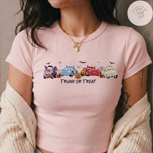 May include: A light pink short-sleeved t-shirt featuring a graphic of four cartoon cars dressed in Halloween costumes, including a vampire, a ghost, and a mummy, alongside pumpkins and the text TRUNK OR TREAT printed in black lettering.