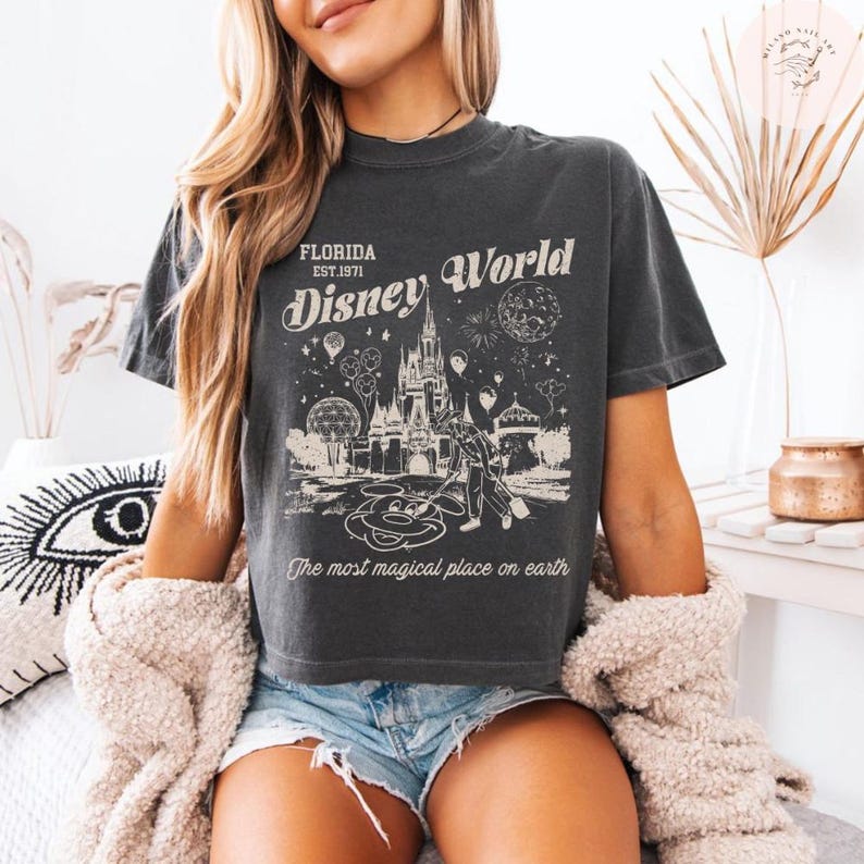 Może przedstawiać: Ciemnoszara koszulka z nadrukiem w stylu vintage przedstawiającym Disney World na Florydzie. Projekt zawiera zamek, Myszka Miki i napis "The most magical place on earth."