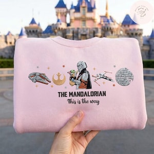 May include: A light pink sweatshirt featuring illustrations of characters and symbols from The Mandalorian. The design includes the Millennium Falcon, a Rebel Alliance symbol, and the text "THE MANDALORIAN This is the way."