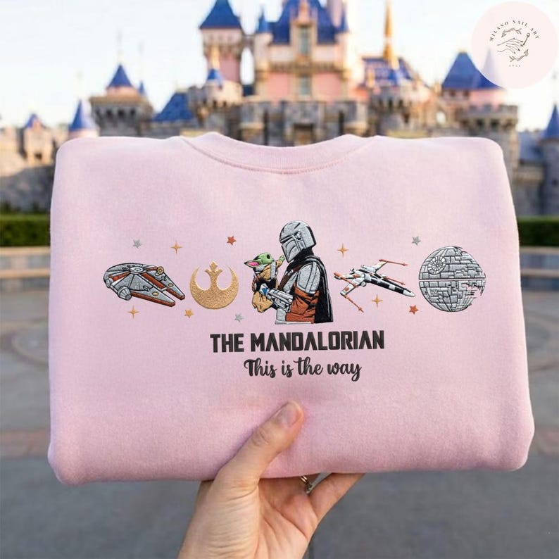 May include: A light pink sweatshirt featuring illustrations of characters and symbols from The Mandalorian. The design includes the Millennium Falcon, a Rebel Alliance symbol, and the text "THE MANDALORIAN This is the way."