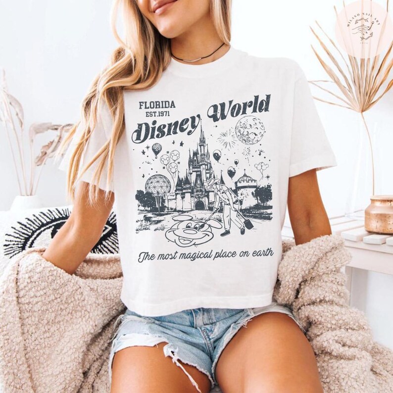 Może przedstawiać: Biała koszulka z czarną grafiką zamku Disney World na Florydzie, fajerwerkami i Myszka Miki. Tekst na koszulce brzmi "FLORIDA EST.1971 Disney World" i "The most magical place on earth."