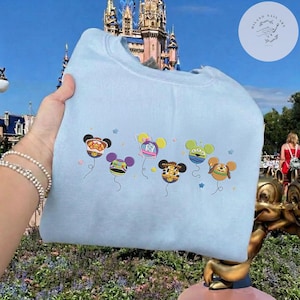 May include: A light blue sweatshirt featuring embroidered Mickey Mouse-themed balloon designs. The balloons are in various colors and patterns, with star accents. The background shows a castle and a park setting.