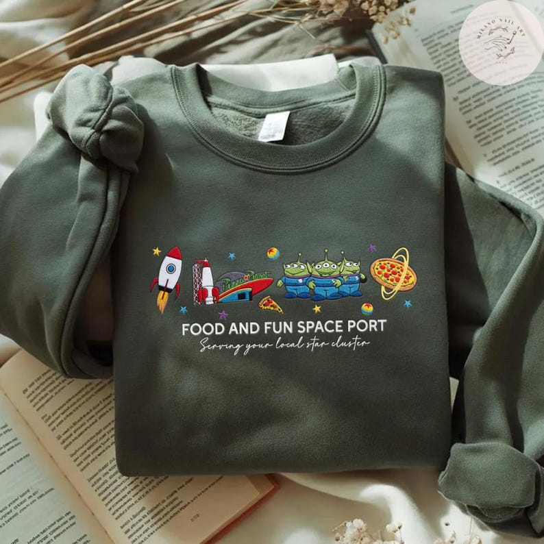 May include: Olive green sweatshirt with a colorful graphic of a rocket, aliens, and pizza. The text "FOOD AND FUN SPACE PORT" and "Serving your local star cluster" are printed below the graphic.
