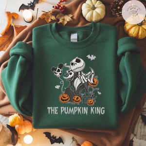 May include: A dark green crewneck sweatshirt featuring a graphic of a skeletal character with a white skull face, wearing a pinstripe suit, surrounded by orange pumpkins, a skull-shaped balloon, and bats, with the text THE PUMPKIN KING below.