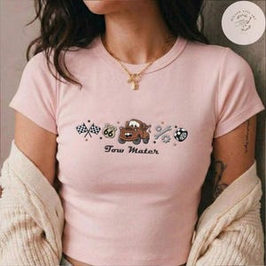 May include: A light pink short-sleeved t-shirt featuring a central graphic of a brown tow truck character, a Route 66 sign, checkered flags, a wrench, a gear, and a tire. The text Tow Mater is printed below the graphic in a dark brown font.