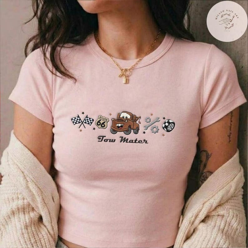 May include: A light pink short-sleeved t-shirt featuring a central graphic of a brown tow truck character, a Route 66 sign, checkered flags, a wrench, a gear, and a tire. The text Tow Mater is printed below the graphic in a dark brown font.