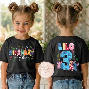 May include: Black t-shirts with colorful graphics. The front of the shirt says "Birthday girl" with a tropical theme. The back of the shirt says "Lilo 3" with cartoon characters. The girl is wearing blue jeans.