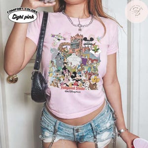 Puede incluir: Camiseta rosa claro Comfort Colors con un gr&aacute;fico colorido de personajes de Disney y el texto "Hollywood Studio, Walt Disney World". La camiseta tiene cuello redondo y mangas cortas. El texto "Light pink" est&aacute; en un &oacute;valo blanco en la parte superior izquierda.