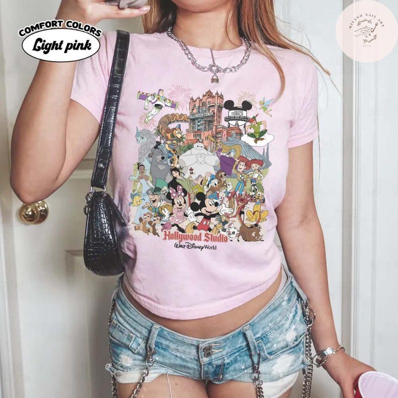 Puede incluir: Camiseta rosa claro Comfort Colors con un gr&aacute;fico colorido de personajes de Disney y el texto "Hollywood Studio, Walt Disney World". La camiseta tiene cuello redondo y mangas cortas. El texto "Light pink" est&aacute; en un &oacute;valo blanco en la parte superior izquierda.