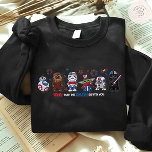 Op de afbeelding: Zwarte crewneck sweatshirt met een patriottisch Star Wars-ontwerp. De afbeelding toont BB-8, Chewbacca, een Stormtrooper, Grogu, R2-D2 en Darth Vader in rood, wit en blauw. Tekst luidt "USA - May the 250th Be With You."