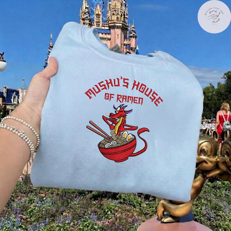 Op de afbeelding: Lichtblauw sweatshirt met de rode tekst "Mushu's House of Ramen". Het ontwerp toont een rode draak met eetstokjes boven een kom ramen. Op de achtergrond zijn een kasteel en een blauwe lucht te zien.