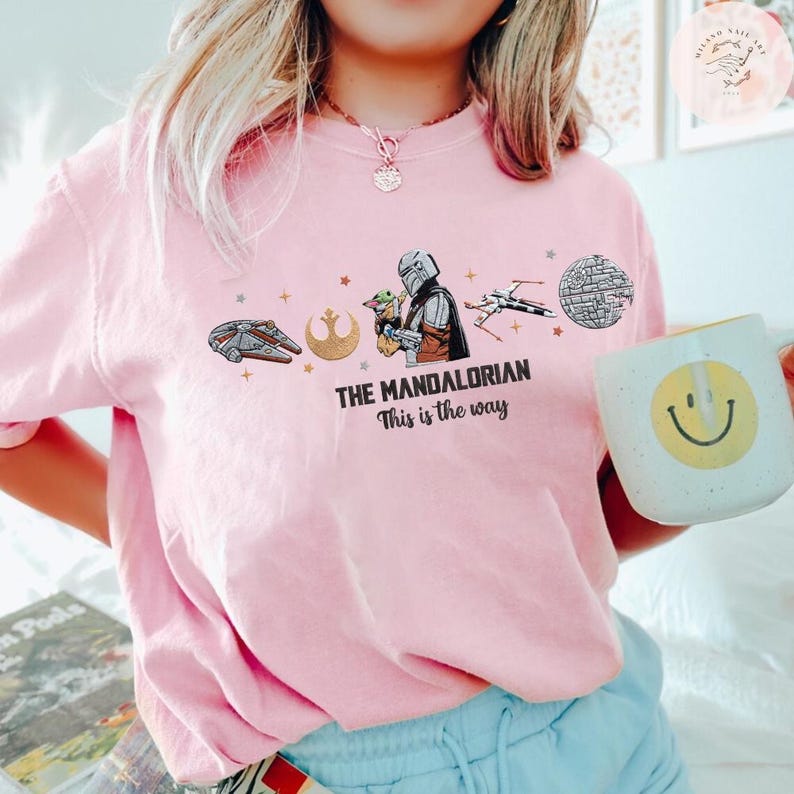 May include: A light pink t-shirt featuring a graphic from "The Mandalorian." The design includes the text "The Mandalorian" and "This is the way," along with illustrations of the Millennium Falcon, the Rebel Alliance symbol, the Mandalorian, Grogu, a TIE fighter, and the Death Star.