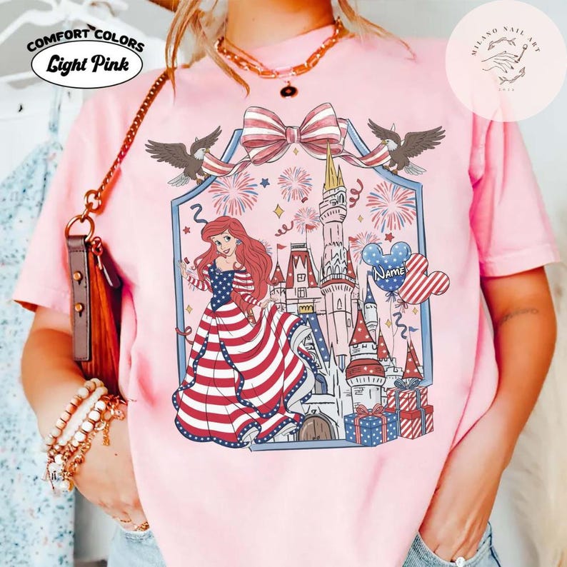 May include: Pale pink Comfort Colors t-shirt with a patriotic design. The graphic features a princess in a red, white, and blue dress, a castle, fireworks, and a bow. The text "Light Pink" is above the design.