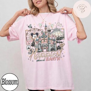 May include: A light pink t-shirt featuring a colorful graphic of a castle, a Ferris wheel, and the text "The Happiest Place on Earth." The design includes Mickey Mouse and other Disney-themed elements.