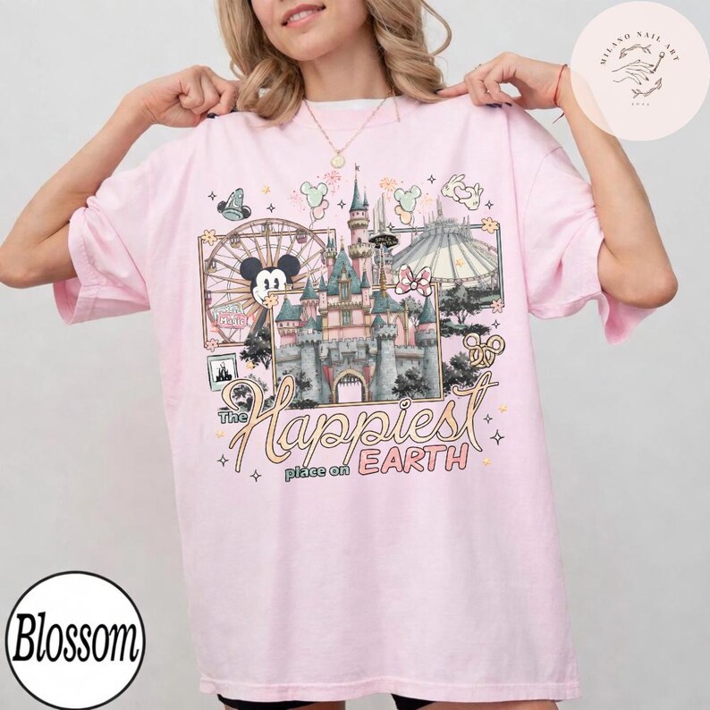 May include: A light pink t-shirt featuring a colorful graphic of a castle, a Ferris wheel, and the text "The Happiest Place on Earth." The design includes Mickey Mouse and other Disney-themed elements.