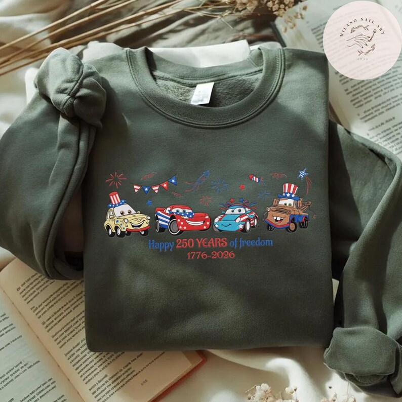 Pode incluir: Um moletom verde-oliva com um gr&aacute;fico de carros de desenho animado em vermelho, branco e azul, decorados com temas patri&oacute;ticos. O texto "Happy 250 Years of freedom 1776-2026" est&aacute; abaixo dos carros. O moletom tem gola redonda e mangas compridas.