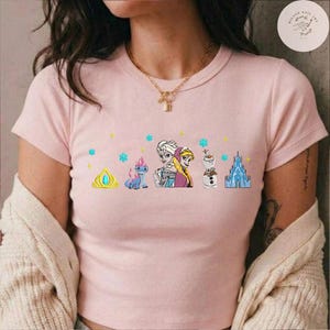 May include: A light pink t-shirt featuring a colorful graphic of characters and elements from the animated movie Frozen. The design includes Elsa, Anna, Olaf, a castle, and other elements. The shirt has short sleeves and a crew neck.