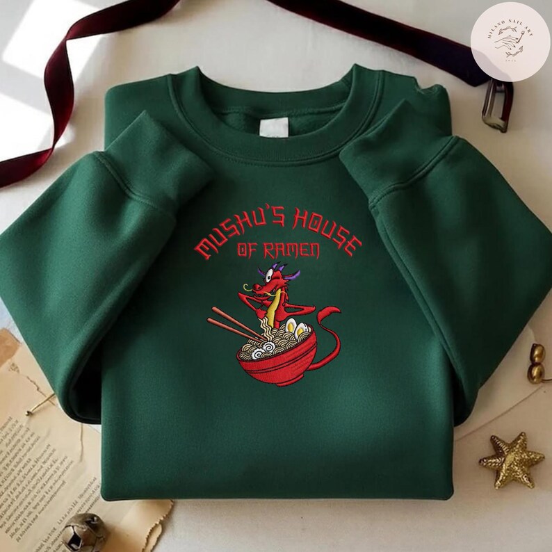 Op de afbeelding: Een donkergroene sweatshirt met de tekst "Mushu's House of Ramen" in het rood. Het ontwerp bevat een geborduurd beeld van een rode draak die ramen eet uit een rode kom, met eetstokjes. De sweatshirt heeft lange mouwen.