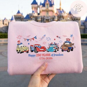 Pode incluir: Camisola rosa claro com um design patri&oacute;tico. Apresenta carros de desenho animado, fogos de artif&iacute;cio e o texto "Happy 250 Years of freedom 1776-2026". O design utiliza as cores vermelho, branco e azul.