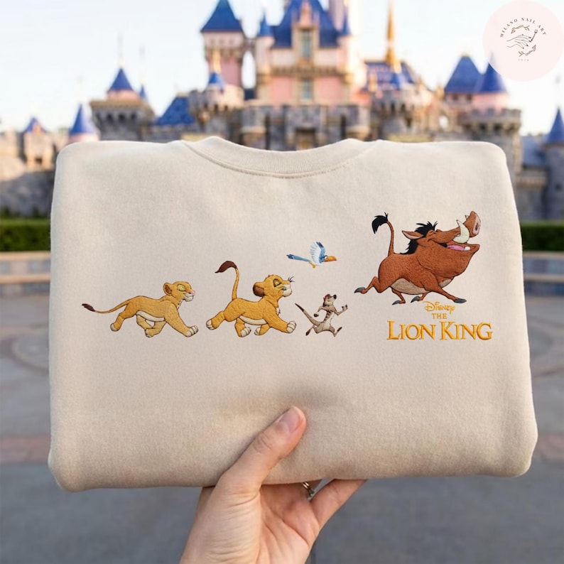 May include: A cream-coloured sweatshirt with an embroidered design from Disney's The Lion King. The design features Simba, Timon, Pumbaa, and a bird, with the words "The Lion King" in gold lettering. The sweatshirt is held by a person.