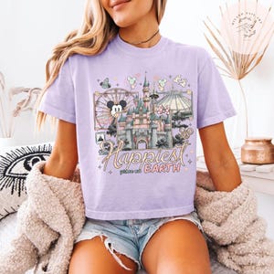 May include: A light purple t-shirt featuring a colorful graphic of a castle, a Ferris wheel with Mickey Mouse, and the text "The Happiest Place on Earth." The design includes other park icons and pastel colors.
