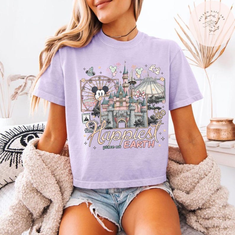 May include: A light purple t-shirt featuring a colorful graphic of a castle, a Ferris wheel with Mickey Mouse, and the text "The Happiest Place on Earth." The design includes other park icons and pastel colors.