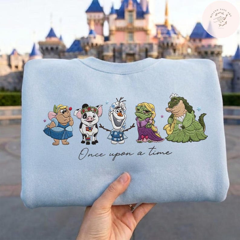 May include: A light blue sweatshirt featuring embroidered cartoon characters, including a mouse, pig, snowman, frog, and alligator. The phrase "Once upon a time" is written below the characters. The sweatshirt is held up in front of a castle.
