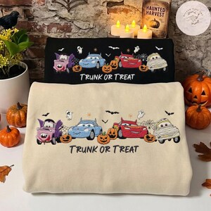 May include: Two sweatshirts, one black and one beige, featuring embroidered cartoon cars dressed in Halloween costumes, including a vampire, a mummy, and a devil, alongside pumpkins, ghosts, and bats, with the text TRUNK OR TREAT below.