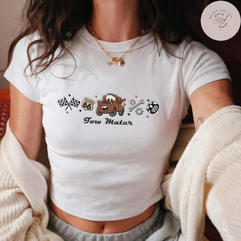May include: A white short-sleeved t-shirt featuring a graphic print of a brown tow truck, a Route 66 sign, checkered flags, a wrench, and a tire. The text "Tow Mater" is printed in black below the graphic. The shirt has a crew neckline.