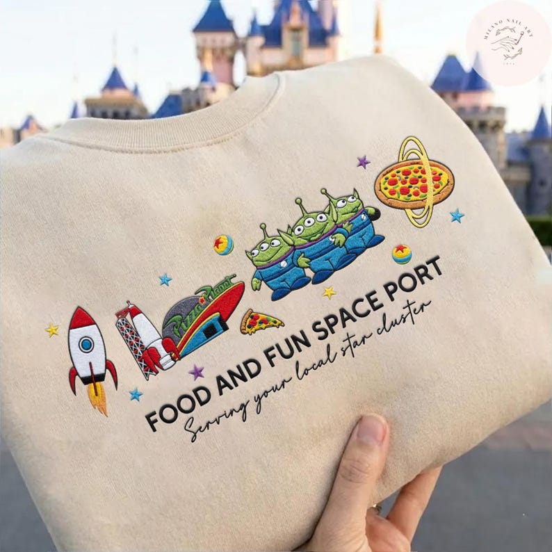 May include: Beige sweatshirt with a colorful embroidered design featuring three green aliens in blue jumpsuits, a rocket ship, pizza, and the text "FOOD AND FUN SPACE PORT". The design includes stars and planets.