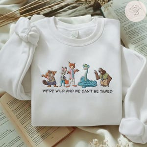 May include: White sweatshirt featuring a colorful cartoon graphic of several animals, including a beaver, rabbit, fox, snake, and sloth. The text "WE'RE WILD AND WE CAN'T BE TAMED" is printed below the graphic. The sweatshirt has long sleeves.
