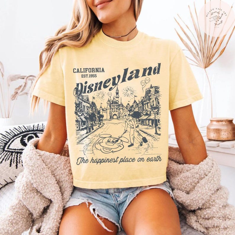May include: Yellow t-shirt with a Disneyland graphic. The graphic features an illustration of a street scene with Mickey Mouse, fireworks, and the text "Disneyland California Est. 1955" and "The happiest place on earth."