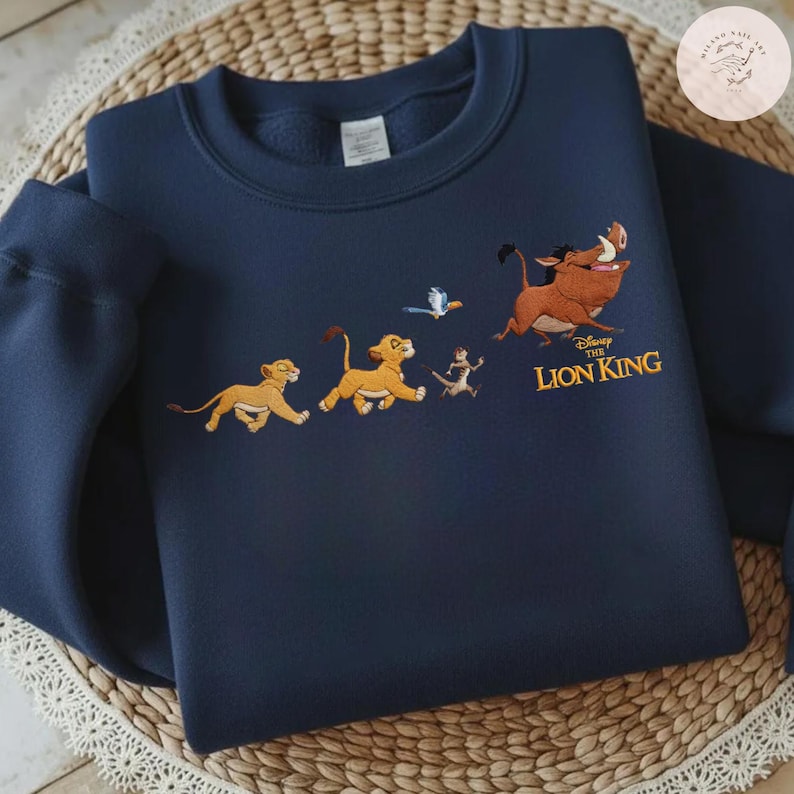 May include: Navy blue sweatshirt with an embroidered design from Disney's The Lion King. The design features Simba, Timon, Pumbaa, and Zazu running, with the film's title in gold lettering.