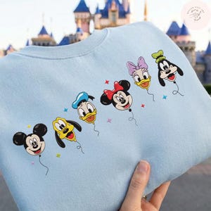 May include: Light blue sweatshirt featuring embroidered Disney characters as balloons. Mickey Mouse, Pluto, Donald Duck, Minnie Mouse, Daisy Duck, and Goofy are depicted as colorful balloons with star accents. A fun and whimsical design.